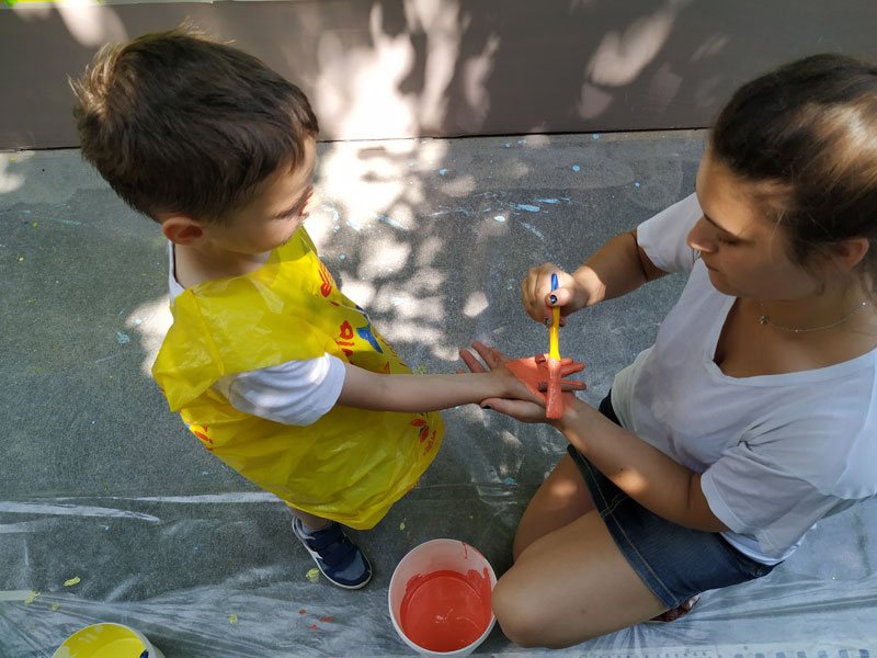 L'immagine ritrae un bambino della scuola dell'infanzia mentre un'alunna del Liceo Artistico gli dipinge la mano per la realizzazione del murales
